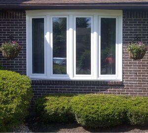 A white bay window set in a dark brick wall, flanked by two hanging flower baskets. Neatly trimmed green bushes are planted below the window.