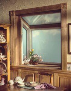 A cozy kitchen nook with wooden trim features a bright window sill decorated with a yellow tulip, potted plants, and a basket. A table below holds a floral teapot set, a plate, and a folded lavender napkin.