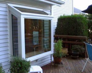 A bay window extends from the white siding of a house onto a wooden deck, surrounded by various potted plants and greenery. A white stool sits near the window, and a blue patio chair is partially visible.