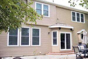 A two-story beige house with large windows and patio doors Denver opens to a patio with outdoor furniture and an umbrella. Green trees partially frame the view.