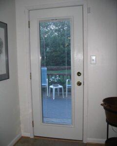 A white door with a tall, narrow window pane and closed blinds. Outside, two white chairs and a small table with a potted plant are visible on a wooden deck surrounded by greenery.