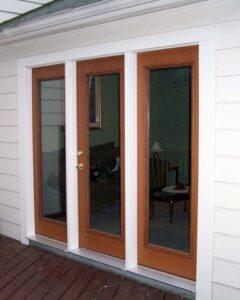A set of wooden-framed glass French doors with a narrow glass panel between them, set in a white exterior wall, leading to a room with a chair, lamp, and table visible inside.