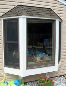 A bay window with white trim on a beige house exterior. The window reflects outdoor furniture and contains a potted plant and decorative items on the inside ledge. Multicolored balls and flowers are near the base outside.