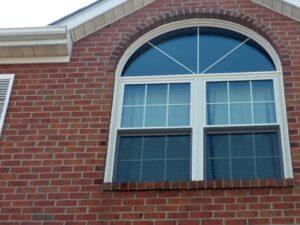 A close-up of a red brick house facade shows a large window with a semi-circular arch on top and rectangular panes below, framed in white. The roof and gutter are partially visible.