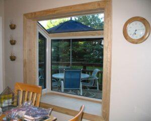 A large bay window with wood trim looks out onto a deck with a round table, blue chairs, and a blue umbrella. Trees are visible outside. A clock hangs on the beige wall inside, and part of a dining table is visible.