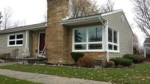 A single-story house with tan siding and a large bay window, stone chimney, red front door, and neatly trimmed shrubs, with a lawn and sidewalk in front. Trees with sparse autumn leaves are in the background.