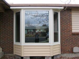 A large bay window with white trim and beige siding protrudes from a brick house. The window reflects trees, a person taking a photo, and houses across the street. The base rests on cinder blocks.