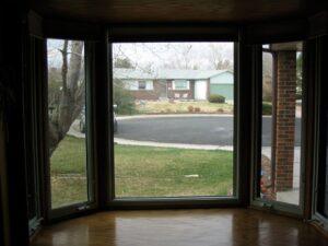 View through a large bay window with three glass panes, looking out at a suburban street, a driveway, and a single-story brick house with a yard and trees on a cloudy day.