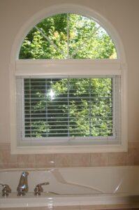 A white-framed window with blinds partially open is above a bathtub with silver faucet handles. Green trees and sunlight are visible through the window, and the top part of the window is arched.