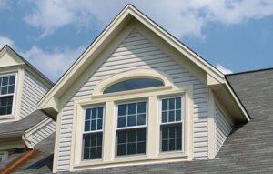 White house gable with cream trim featuring double-hung windows topped by an arched transom, gray shingled roof, and blue sky with clouds in the background.