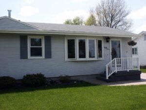A single-story house with light gray siding, white trim, and dark shutters. There is a front porch with white railings, steps leading up, and a hanging flower basket. Green lawn and shrubs are in front.