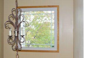 A decorative, metal-framed chandelier with unlit candle-shaped bulbs hangs near a window with a wooden frame, through which green tree branches are visible outside. The interior wall is painted beige.