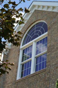 A close-up view of a brick house featuring a large arched window with white trim and divided panes, reflecting blue sky and clouds, partially framed by tree branches and leaves.