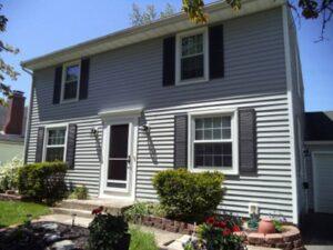 A two-story house with gray siding, black shutters, and white trim, featuring a central front door with small bushes and flowers in front, set on a sunny day.