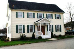 A two-story cream-colored house with green shutters, a black roof, and a white columned front porch sits on a green lawn next to a street. Small shrubs and plants line the walkway to the front door.