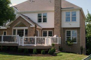 A two-story brick house with white trim, large windows, and a raised deck featuring white railings. The deck has outdoor furniture and is surrounded by neatly landscaped grass and shrubs.