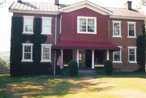 A two-story brick house with white-trimmed windows, ivy growing on part of the left side, a red awning over the porch, and an American flag by the front entrance. Grass lawn is visible in front.