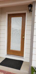 A wooden front door with a large glass panel and mini blinds is set in a light-colored exterior wall. A black doormat lies on the concrete porch, and a wall-mounted lantern is above the door.