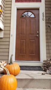 A brown front door with a semicircular window, house number 690, and two orange pumpkins on the steps, suggesting autumn or Halloween decor. Logs and a wind chime are nearby.