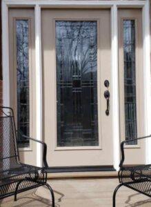 Beige front door with decorative glass panel and matching glass side panels, flanked by two black metal chairs on a porch. Trees and sky are reflected in the glass.