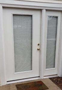 A white exterior door with a large glass panel and matching sidelights, all featuring closed blinds. Two brass door handles are visible above a worn brown doormat on a concrete doorstep.