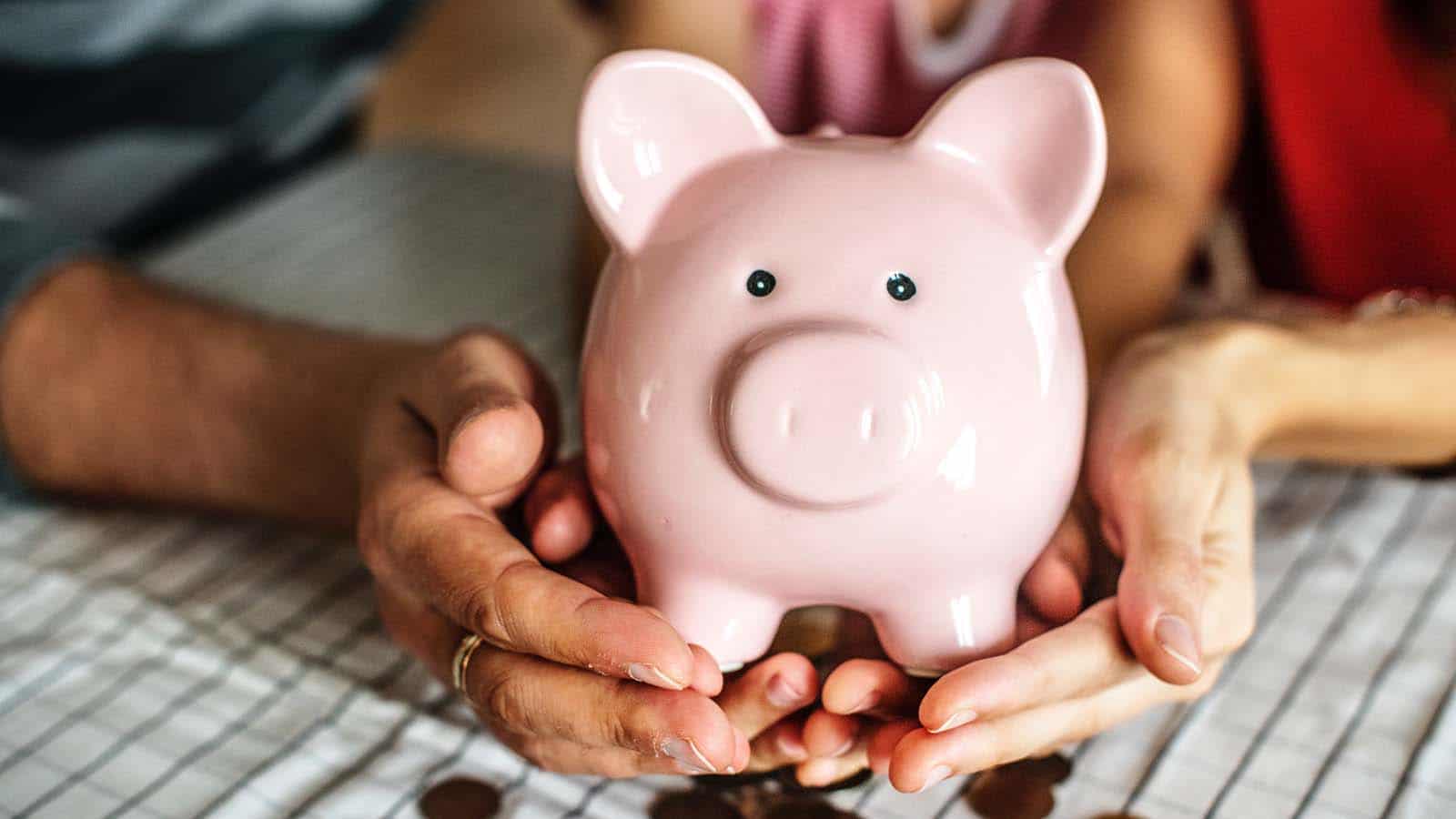 Two people gently hold a pink piggy bank together on a table with scattered coins, symbolizing saving money or financial planning.