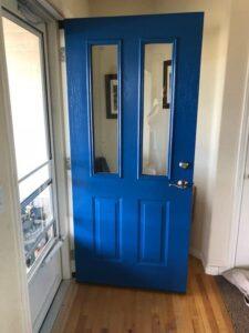 A bright blue front door is installed backwards in the doorway, with its handle and locks on the inside edge, making it impossible to close properly. Sunlight streams through nearby windows onto the wooden floor.