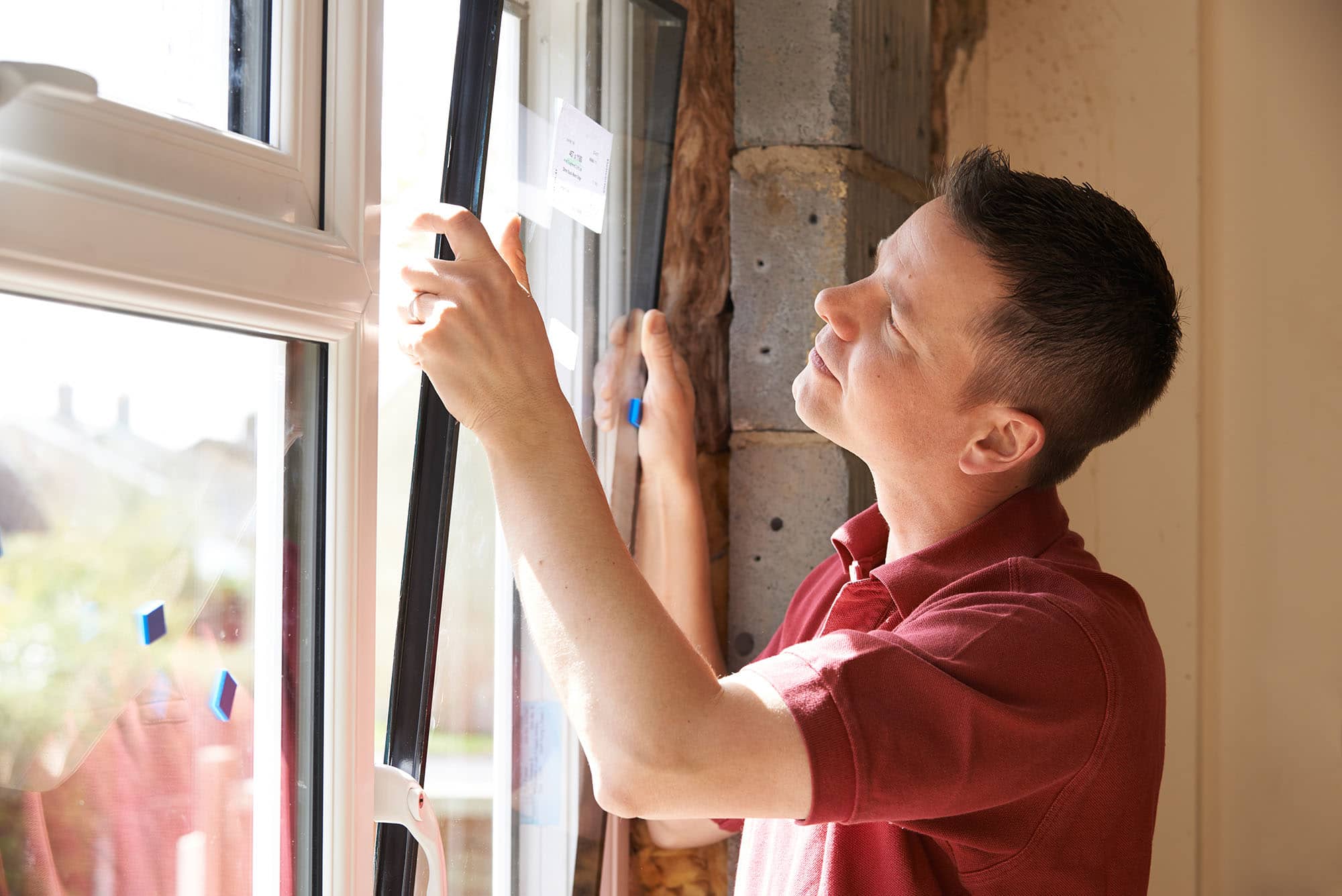 A man in a red polo shirt installs or inspects a window, holding a measuring tape and checking the frame. Sunlight comes through the window, illuminating the workspace.