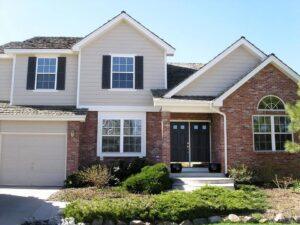 A modern suburban house with beige siding, red brick accents, black shutters, a double front door, attached garage, front steps, and landscaped bushes and plants in the yard.