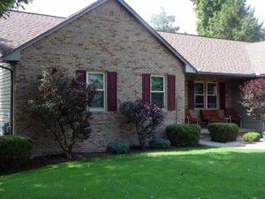 A single-story brick house with two front windows featuring maroon shutters, a covered porch with chairs, neatly trimmed bushes, small trees, and a well-maintained green lawn.