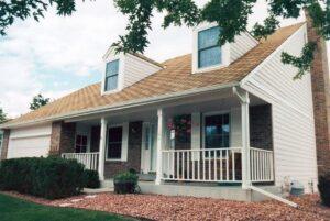 A two-story suburban house with a beige roof, white siding, brick accents, a front porch with white railings, hanging flowers, and a well-kept yard with shrubs and decorative rocks.