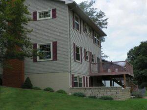A beige two-story house with maroon shutters, a brick chimney, and an elevated wooden deck. The house is surrounded by green grass, small shrubs, and trees. A retaining wall supports the deck area.