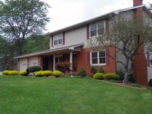 A two-story suburban house with tan siding, red brick accents, and red shutters. The front yard is landscaped with shrubs, small trees, and a well-maintained green lawn.