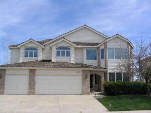Two-story suburban house with white siding, brick accents, three-car garage, multiple front-facing windows, and a neatly trimmed lawn and shrubs under a blue sky.
