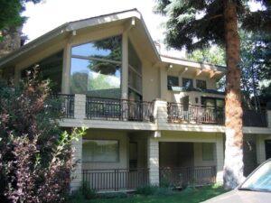 A two-story modern house with large, angled windows, beige siding, and a spacious balcony. Surrounded by trees and greenery, the home has a sloped roof and dark railings on both levels.