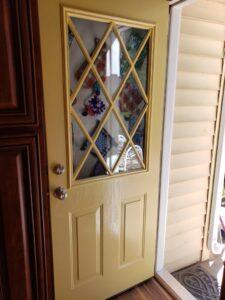 A yellow door with diamond-patterned window panes is slightly open, revealing sunlight and the beige siding outside. Inside, kitchen cabinets and colorful pot holders are visible.
