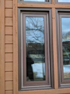 A tall, narrow window with brown trim on a wooden exterior wall reflects bare tree branches, a building, and patches of snow on the ground outside.