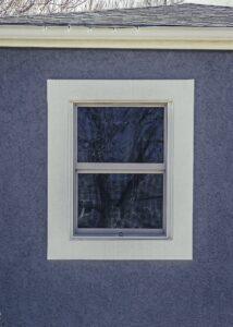 A single rectangular window with a white frame is set in a textured gray wall. Bare tree branches are reflected in the window glass. The roof edge and some string lights are visible at the top.