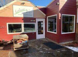 A small red building with white trim, large windows, and a sign above the door that reads Artisan You: Creative Studio. A wooden cart stands outside on a partially wet sidewalk under a clear sky.
