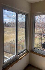 Two large windows in a corner let in sunlight and offer a view of a fenced backyard, trees with bare branches, and a grassy field with houses in the distance under a partly cloudy sky.