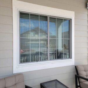 A large rectangular window with white trim reflects a house and sky. Two cushioned chairs and a glass-top table sit on a gray-sided porch in front of the window.