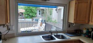 A kitchen sink with a double basin is set in a light countertop below a large window, which looks out onto a patio with white lounge chairs and a view of neighboring houses and greenery.