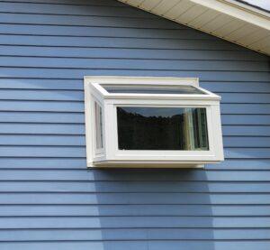 A white-framed greenhouse window protrudes from the side of a blue house with horizontal siding, allowing extra light and space inside.