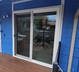 A white-framed sliding glass door on a blue-paneled house opens to a wooden deck with a table and chairs, partially covered by a closed red patio umbrella. A house and trees are visible in the background.