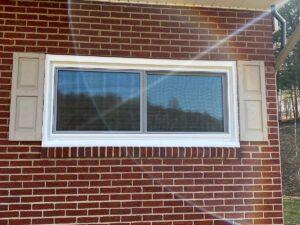 A rectangular window with beige shutters on a red brick wall, reflecting sunlight and a faint rainbow lens flare across the image. Trees and a hill are visible faintly through the window.