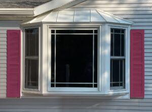 A white bay window with three sections and a metal roof, framed by bright pink shutters, set in the light-colored exterior wall of a house.