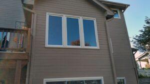 A beige house exterior with a set of three large, rectangular windows on the second floor, reflecting the sky and trees. A wooden deck is on the left, and part of a white fence and tree are visible to the right.