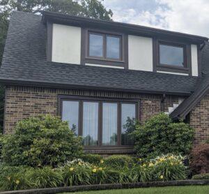 A brick house features a dark gray shingled roof, three large windows on the lower level, two windows on the upper level, and neatly trimmed bushes and yellow flowers in the front yard.