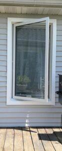 A white-framed window on a light gray house exterior is open outward. Reflections of plants and curtains are visible in the glass. A black grill is partially visible to the right on a wooden deck.
