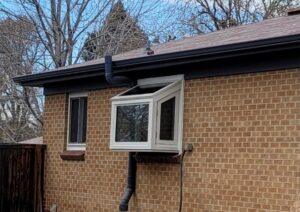 A brick house with an unusual window installation; the window is tilted outward and fitted with black drainage pipes extending from the roof and window down the exterior wall. Leafless trees are visible in the background.